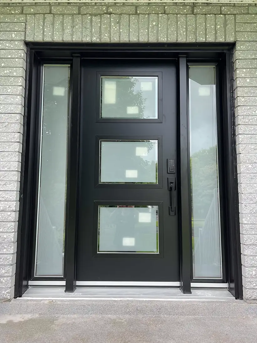 Porte d'entrée noire vitrée à trois panneaux avec deux panneaux latéraux givrés, sur brique claire.