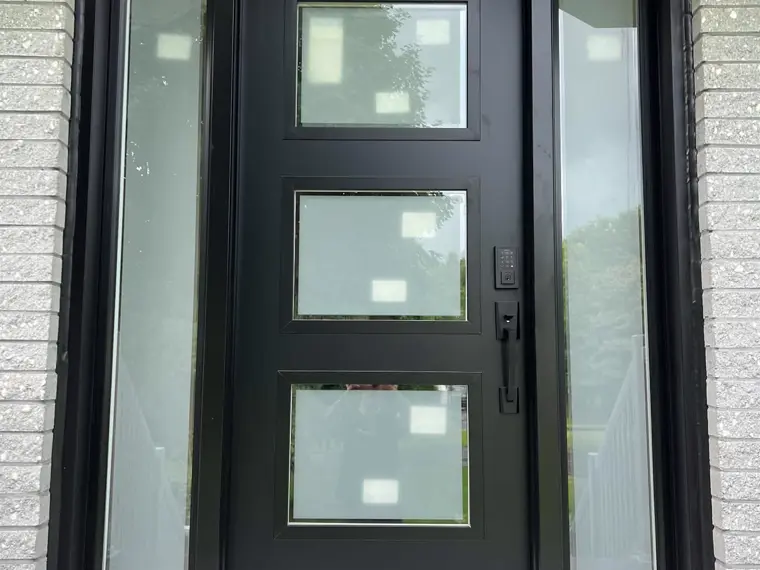 Porte d'entrée noire vitrée avec panneaux latéraux, sur brique blanche.